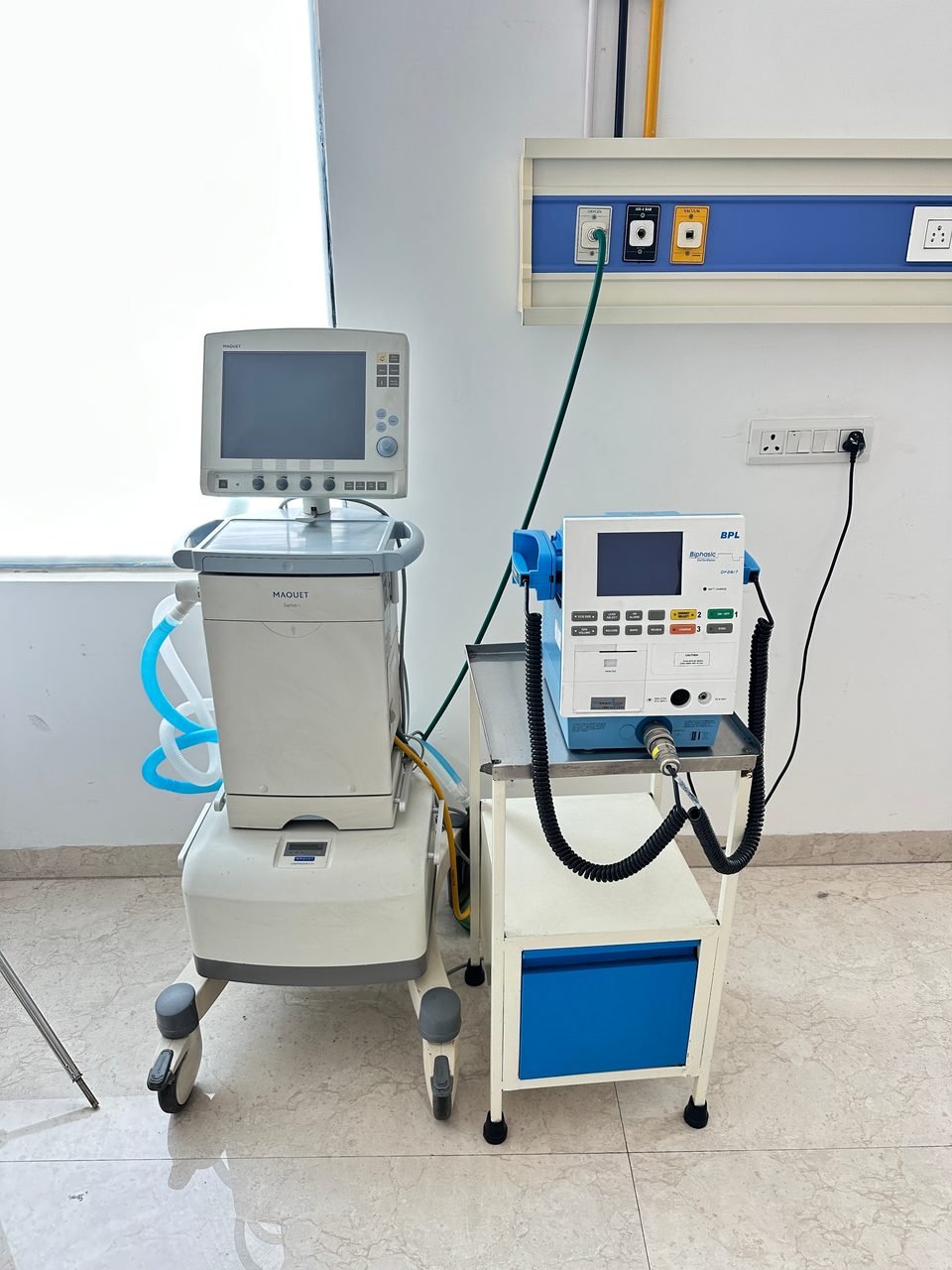 A ventilator and a defibrillator on trolleys beside a medical gas panel